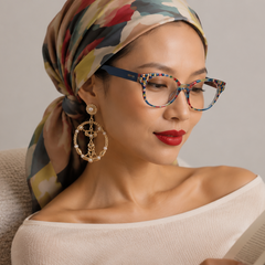 A woman wearing a multicolor patterned headscarf and blue patterned cat-eye glasses, reading a book while wearing large gold hoop earrings, set against a neutral gray background.